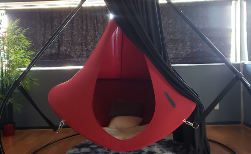 A red cocoon tent suspended from a black frame. It has a black curtain drawn over it to create a secluded space.